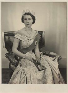 A sepia-toned photograph of a young Queen Elizabeth wearing a crown, a dress and a sash, sitting on an armchair