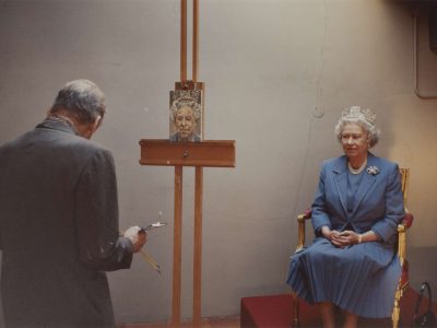 A photo of an artist (Lucian Freud) with his back to the viewer, in front of an easel. An older lady (Queen Elizabeth II) sits in front of the easel, wearing a blue suit.