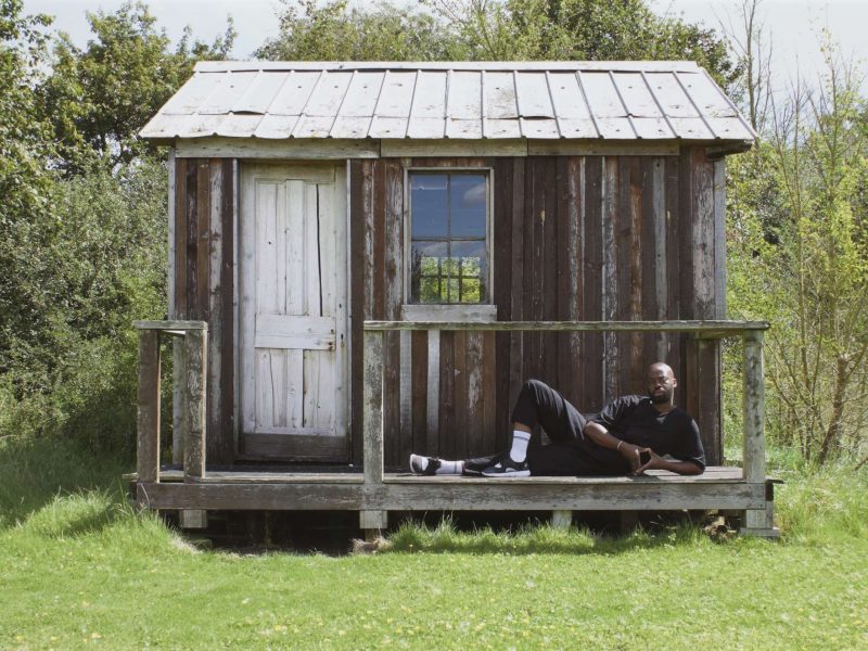 A small, wooden cabin on green grass with trees behind it; a man wearing all black lounges at its step.