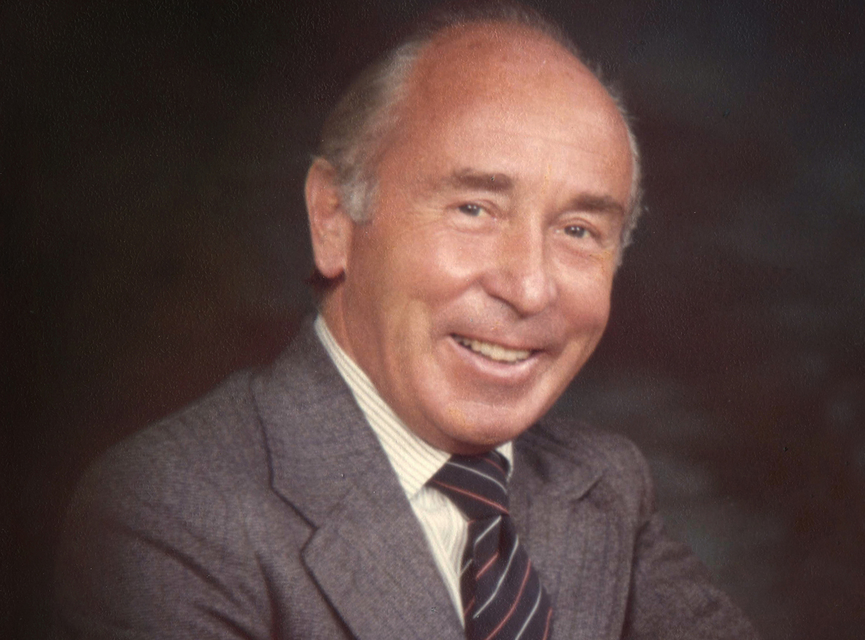 A photo of a man with his arms crossed in front of his chest. He is wearing a grey suit and a dark grey tie, and is smiling at the camera.
