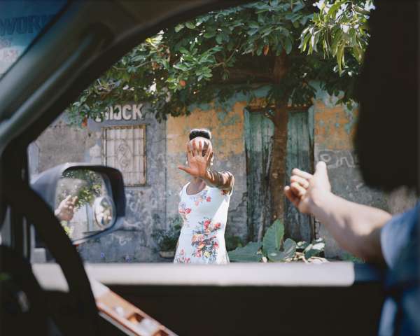 A photo taken from inside a car; a man in a shirt reaches his arm out and a black woman outside, perfectly framed in the car window, reaches her arm, her hand covering her face.