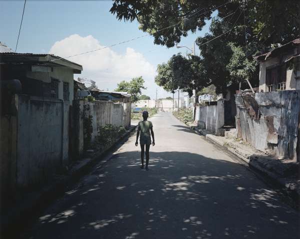 A small child in shorts has its back turned to the camera; the cild is walking down a street, shady under the shadow of a huge tree.