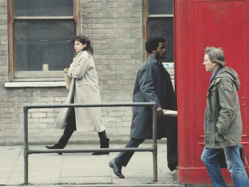Three people walk into the shot; a white man walks past a red phonebox; a black man appears to be walking into the phonebox and a white woman clutches her coat tighly, looking cold.