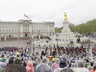 A photo of Buckingham Palace and thousands of people watching as a parade starts in front of them; it's a grey, gloomy day, and everyone is wearing waterproofs